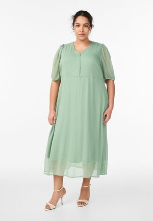 Žena s kučeravými vlasmi v svetlozelenej midi sukni s nafúknutými rukávmi a gombíkmi, v kombinácii s béžovými sandálmi na vysokom podpätku, stojaca.