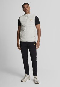 Veste matelassée gris clair avec une fermeture éclair sur le devant et deux poches latérales. Présente un petit logo jaune sur la poitrine. Portée par-dessus un t-shirt noir et un pantalon.