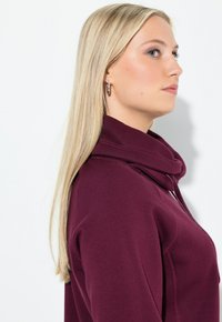 Felpa bordeaux con colletto alto, cuciture in vista e texture morbida, caratterizzata da una vestibilità rilassata e maniche lunghe.