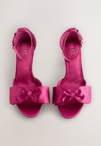 Fuksianväriset satiiniset korkokengät, joissa leveä rusetti edessä ja nilkkahihna. Kengissä on sileä, kiiltävä pinta.