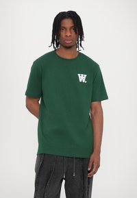 Green cotton t-shirt featuring short sleeves and a round neckline, with a white logo on the left chest area. Smooth texture and casual fit.