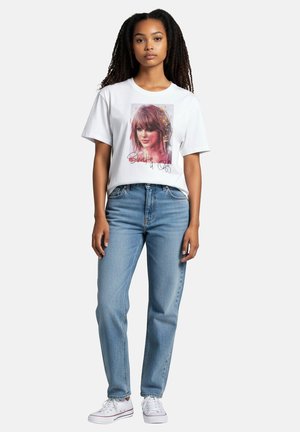 Junge Frau mit langen lockigen Haaren, die ein weißes T-Shirt mit Grafik, blaue Jeans und weiße Turnschuhe trägt und vor einem einfarbigen Hintergrund steht.