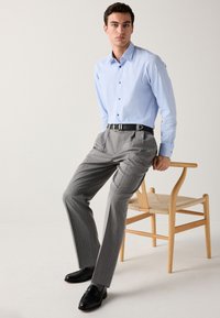 Ljusblå skjorta med knappar, grå skräddarsydda byxor med tunt ränder, svarta läderskor och svart bälte. Sitter på en trästol med flätad sits.