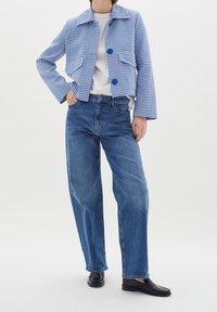 Persona vestida con una chaqueta de cuadros azules sobre una camisa blanca, jeans de tiro alto azules y mocasines negros, de pie con una mano en el bolsillo.