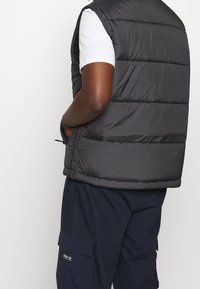 Černá prošívaná vesta s vysokým límcem, bočními kapsami na zip a viditelným horizontálním prošitím. Nositelná přes bílou košili a tmavě modré kalhoty.