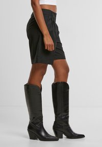Černé kožené boty ke kolenům s špičatou špičkou a blokovým podpatkem, spárované s perfektně střiženými černými kraťasy s předním záhybem a knoflíkovým zapínáním.