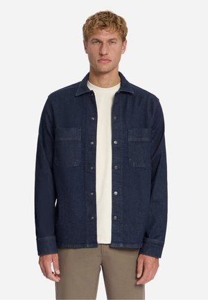Man wearing a dark blue buttoned denim jacket over a white shirt and brown pants, standing against a plain light background.