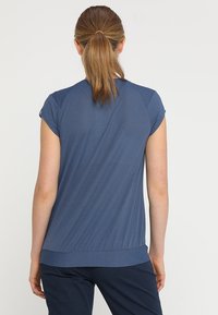 Femme aux cheveux bruns attachés en queue de cheval, portant un haut bleu à manches courtes et un pantalon noir, vue de dos sur un fond uni.