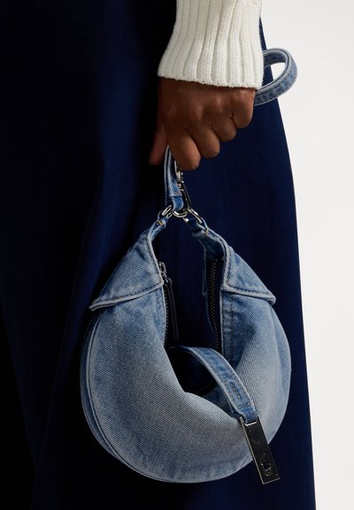 Kerekített, kék farmer anyagból készült kézitáska texturált felülettel, cipzárokkal és fémes pántkapcsolattal, egy kéz által tartva.