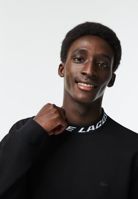 Zwarte sweatshirt met een ronde hals en een opvallende groene en witte merk-kraag. Subtiele logo op de borst, gladde textuur.