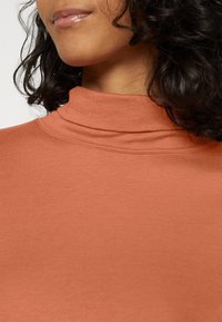 En rostfärgad polokrage i mjukt och stretchigt tyg, med en figurnära design och lagerdetaljer vid halslinningen.