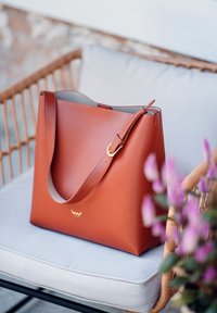 Braune Ledertragetasche mit breitem Körper, verstellbarem Riemen und goldenen Akzenten. Glatte Textur mit minimalistischem Design.