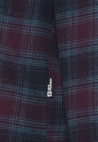 Tessuto a quadri in rosso scuro e blu navy, con una texture morbida e sottili linee blu. Include un'etichetta con il marchio "Jack Wolfskin".