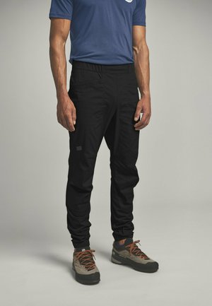 Mann, der schwarze Cargo-Hosen mit elastischen Bündchen und grauen Wanderschuhen mit orangenen Schnürsenkeln trägt, steht vor einem neutralen Hintergrund.