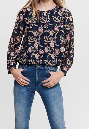 Blusa a maniche lunghe blu navy con motivo floreale in rosa e beige. Tessuto leggero, scollatura rotonda, vestibilità comoda, abbinata a jeans azzurri chiari.