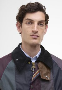 Többszínű kabát, amelyen bordás gallér, arany cipzár és sötét árnyalatú, különböző textúrájú panelek találhatók. Szivárványcsíkkal díszítve.