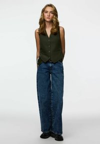 Maslinasto zelena prsluk bez rukava s V-izrezom i gumbima, uparena s širokim plavim trapericama i crnim cipelama. Jednostavan dizajn i ležerni kroj.