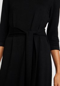 Robe noire avec manches trois-quarts et ceinture en tissu nouée à la taille, portée par une personne au teint moyen.