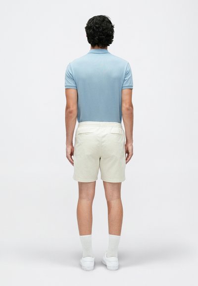 Gai zils polo krekls kopā ar krēmīgām šortiem, kam ir elastīgs josta. Modelis valkā baltas sporta apavus un garās zeķes.