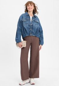 Denim jasje met een cropped ontwerp en twee borstzakken, gecombineerd met brede bruine broeken en lichtgekleurde sneakers. Houdt een kleine crèmekleurige handtas vast.