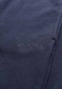 Tejido de algodón azul marino con una textura suave que presenta la palabra bordada "SUPERDRY" en un arco. Se pueden ver los detalles de las costuras a lo largo de los bordes.