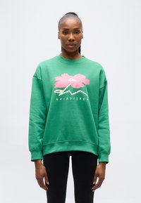 Γυναίκα που φορεί πράσινο φούτερ με ροζ λουλούδι και λευκό λογότυπο, συνδυασμένο με μαύρο κολάν, στέκεται μπροστά σε ένα απλό ανοιχτό φόντο.