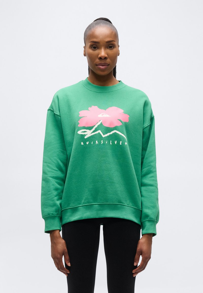 Γυναίκα που φορεί πράσινο φούτερ με ροζ λουλούδι και λευκό λογότυπο, συνδυασμένο με μαύρο κολάν, στέκεται μπροστά σε ένα απλό ανοιχτό φόντο.