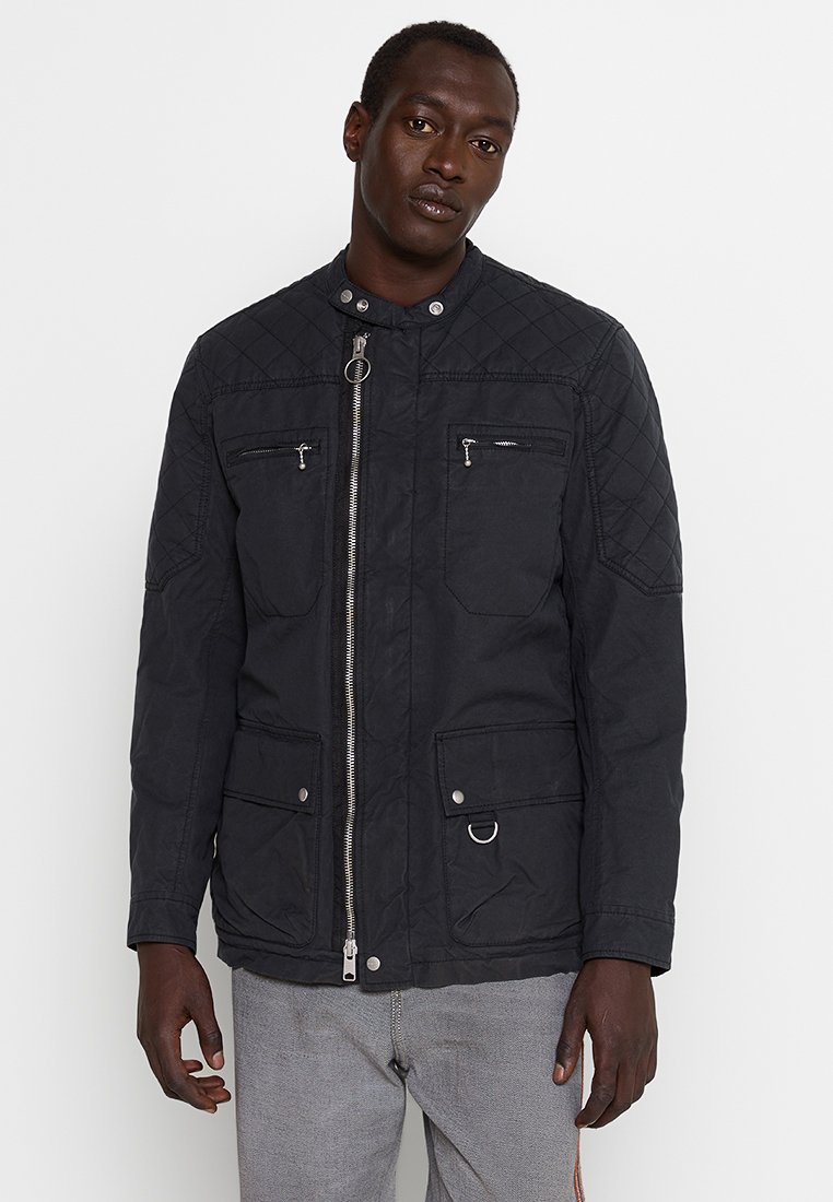 Schwarze gesteppte Jacke mit Stehkragen, silbernem Reißverschluss und mehreren Fronttaschen. Verfügt über auffällige Nähte und subtile Hardware-Details.