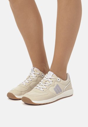 Pies vistiendo zapatillas beige y blancas con cordones, un logo lila claro y suelas de goma sobre un fondo blanco.