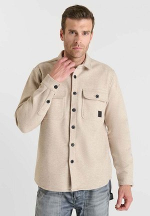 Man draagt een beige overhemdjas met klepzakken, staat voor een effen lichte achtergrond en stelt de kraag bij met zijn linkerhand.