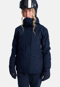 Woman wearing black ski jacket, gloves, and helmet with goggles against a plain background, smiling slightly.