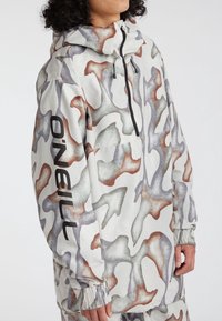 Kapuzende Jacke in einem weißen Grundton mit abstraktem Tarnmuster in gedämpften Farben. Verfügt über elastische Bündchen, eine Fronttasche und einen Reißverschluss.