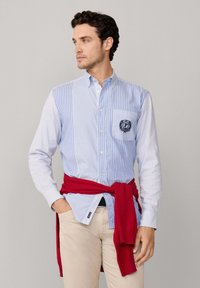 Blauw en wit gestreept overhemd met een zak en logo-detail. Rode trui om de taille geknoopt. Licht beige broek. Glad stof oppervlak.