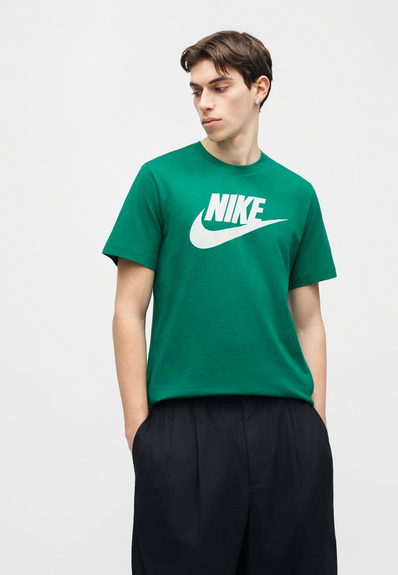 Junger Mann mit kurzem dunklem Haar, der ein grünes Nike-T-Shirt und schwarze Hosen trägt, nach unten und zur Seite schaut, die Hände in den Taschen.