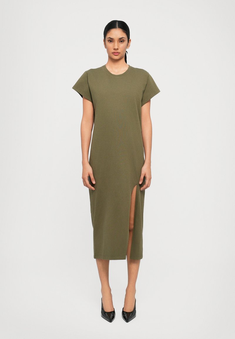 Olive green, knee-length dress with short sleeves and a side slit. Made from soft fabric with a round neckline. Black high-heeled shoes.