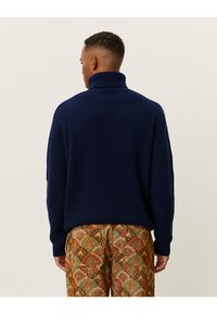 Mornarsko rebrasto pulover s turtleneck ovratnikom in dolgimi rokavi, oblečen preko vzorčastih oranžnih, zelenih in rjavih kordnih hlač, pogled zadaj.