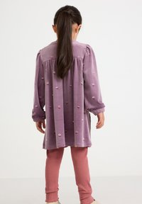 Lila Samtkleid mit Blumenstickerei, langen Ärmeln und elastischen Bündchen, kombiniert mit rosa gerippten Leggings. Einfarbiger Hintergrund.