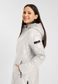 Lichtgrijze, gewatteerde pufferjack met een capuchon; heeft ritszakken en een logopatch op de mouw. Gladde, glanzende textuur.