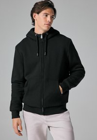 Sort zip-up hoodie med snøre i hætten, forlommer og ribbede manchetter. Glat tekstur og minimalistisk design. Bruges over lyse bukser.