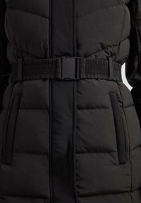 Sort quilted polstret jakke med et bredt elastisk bælte og sort plastiklås i taljen, med to lodrette sidelommer.