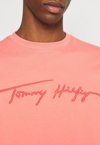 Korala bawełniana koszulka z okrągłym dekoltem i kontrastowym, różowym haftowanym logo "Tommy Hilfiger" na klatce piersiowej. Gładka faktura.