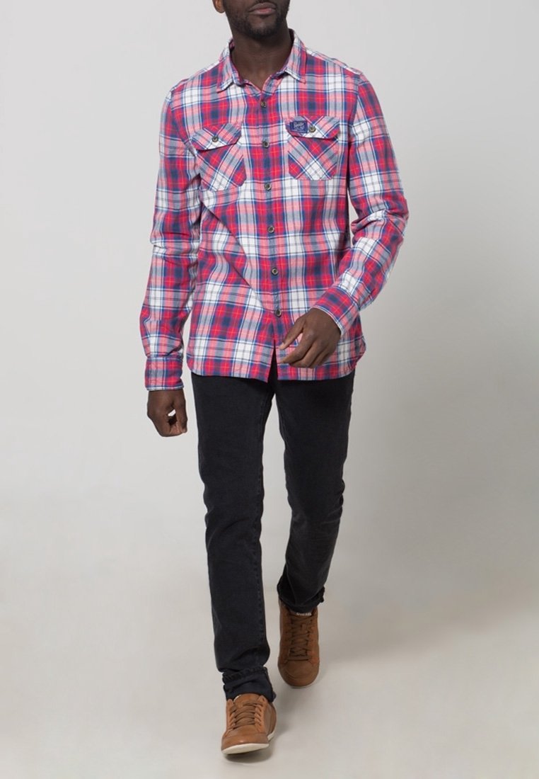Homme marchant portant une chemise à carreaux rouge, blanche et bleue, un pantalon noir et des baskets marron sur un fond clair uni.