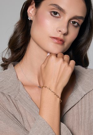 Złota bransoletka z połączonymi owalnymi ogniwami na nadgarstku, w towarzystwie srebrnego naszyjnika. Modelka ma na sobie lekki makijaż i bluzkę w neutralnych odcieniach.