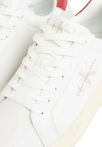 Sneakers bianche in pelle con lacci piatti, lati perforati e un accentuazione rossa sul tallone. Presentano il logo dorato in rilievo: "Calvin Klein Jeans."