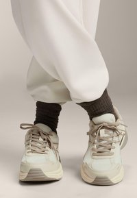 Sneakers beige e bianchi con lacci marroni indossati con calze marroni scure e pantaloni bianchi larghi su uno sfondo neutro.