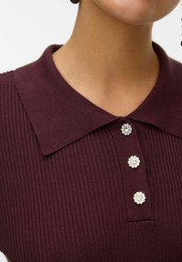 Polo shirt ribbed bordeaux con colletto classico e tre bottoni decorati con design floreali e accenti metallici.