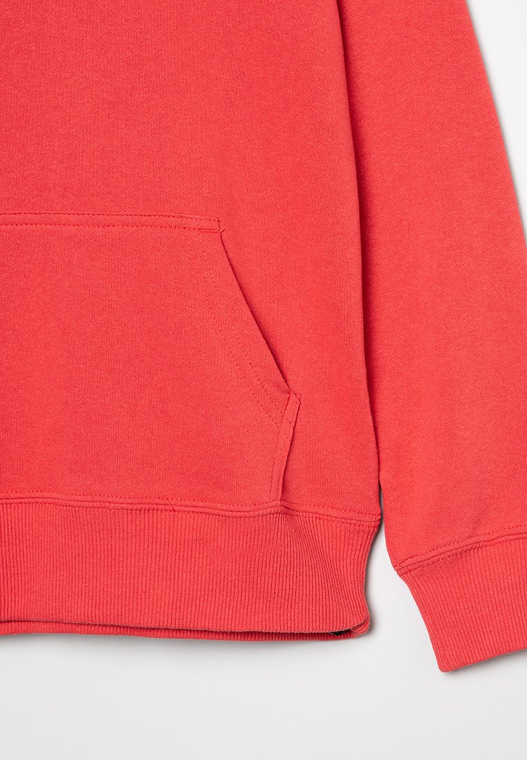 Manga de sudadera roja con puño de canalé y bolsillo tipo canguro sobre un fondo claro liso, que muestra la textura del tejido de punto y los detalles de la costura.