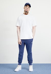 Λευκό βαμβακερό t-shirt και ναυτικές φόρμες, με ελαστικές μανσέτες. Λευκά αθλητικά παπούτσια και ναυτικό καπέλο με λογότυπο. Ουδέτερο φόντο.