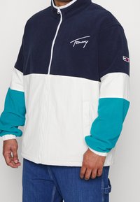 Kurtka Tommy z granatową górną częścią, bia�łym środkiem i zielonkawoniebieską dolną częścią. Posiada wysoki kołnierz, zamek na przodzie i ściągacze na mankietach. Logo na piersi.