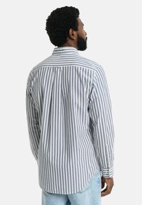 Chemise à rayures à manches longues en blanc et bleu marine avec un col à bouton, un empiècement au dos et un ourlet arrondi ; le tissu a une texture lisse.